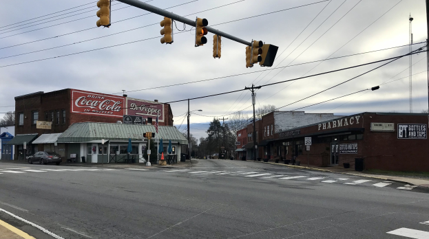 Stadtstraße mit Ampelkreuzung, Fahrzeugen, Gebäuden mit Schildern, Strommasten und Leitungen, Bäumen und bewölktem Himmel.