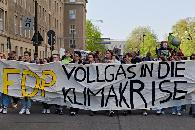 Gruppe von Menschen, die eine Straße entlanggehen und ein Banner mit der Aufschrift "FDP Vollgas in die Klimakrise" halten, mit Gebäuden, Bäumen, Polen, Verkehrszeichen und einem klaren blauen Himmel im Hintergrund.