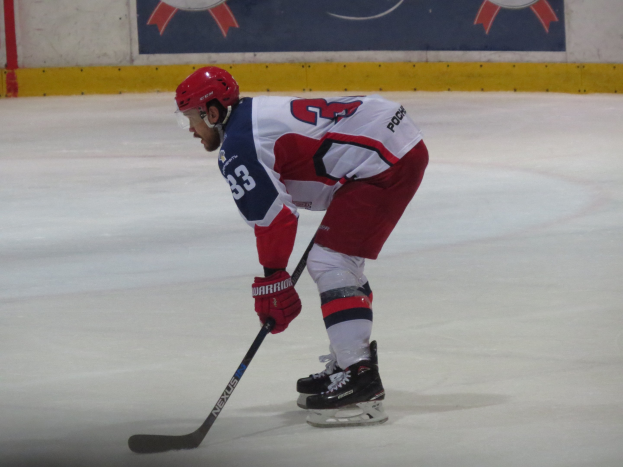Eishockeyspieler in voller Ausrüstung auf dem Eis mit einem Stock, der einer Wand mit einem Banner im Hintergrund gegenübersteht.