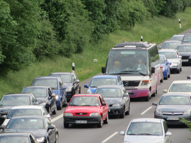 Ein Stau auf einer Autobahn mit vielen Autos und einem Lieferwagen, Menschen in den Fahrzeugen und Bäume und Gras im Hintergrund.
