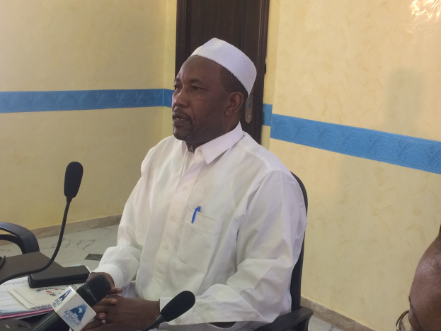 Ein Mann in einem weißen Kleid und einer Mütze sitzt an einem Tisch mit Mikrofonen, spricht, während Papiere und Gegenstände auf dem Tisch sichtbar sind, mit einer anderen Person mit Brille zu seiner Rechten, in einem Raum mit einer Wand und einer Tür im Hintergrund.