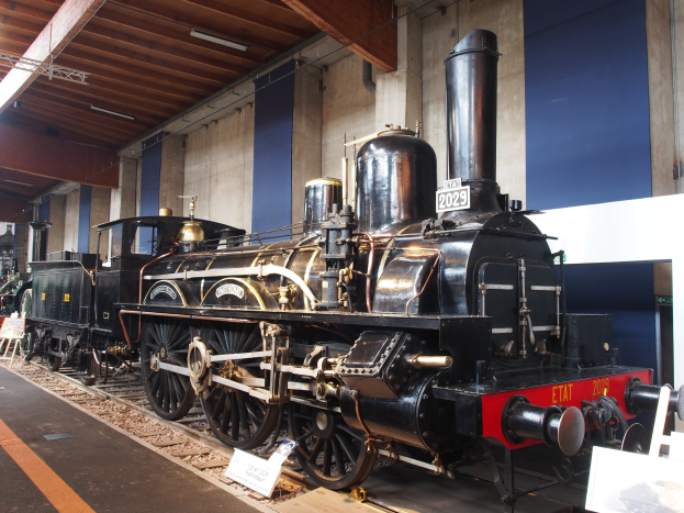Alte Dampflokomotive im Museum ausgestellt, umgeben von Eisenbahnschienen, Informationsschildern, Ständen und anderen Ausstellungsstücken, mit einer Wand und Deckenbeleuchtung im Hintergrund.