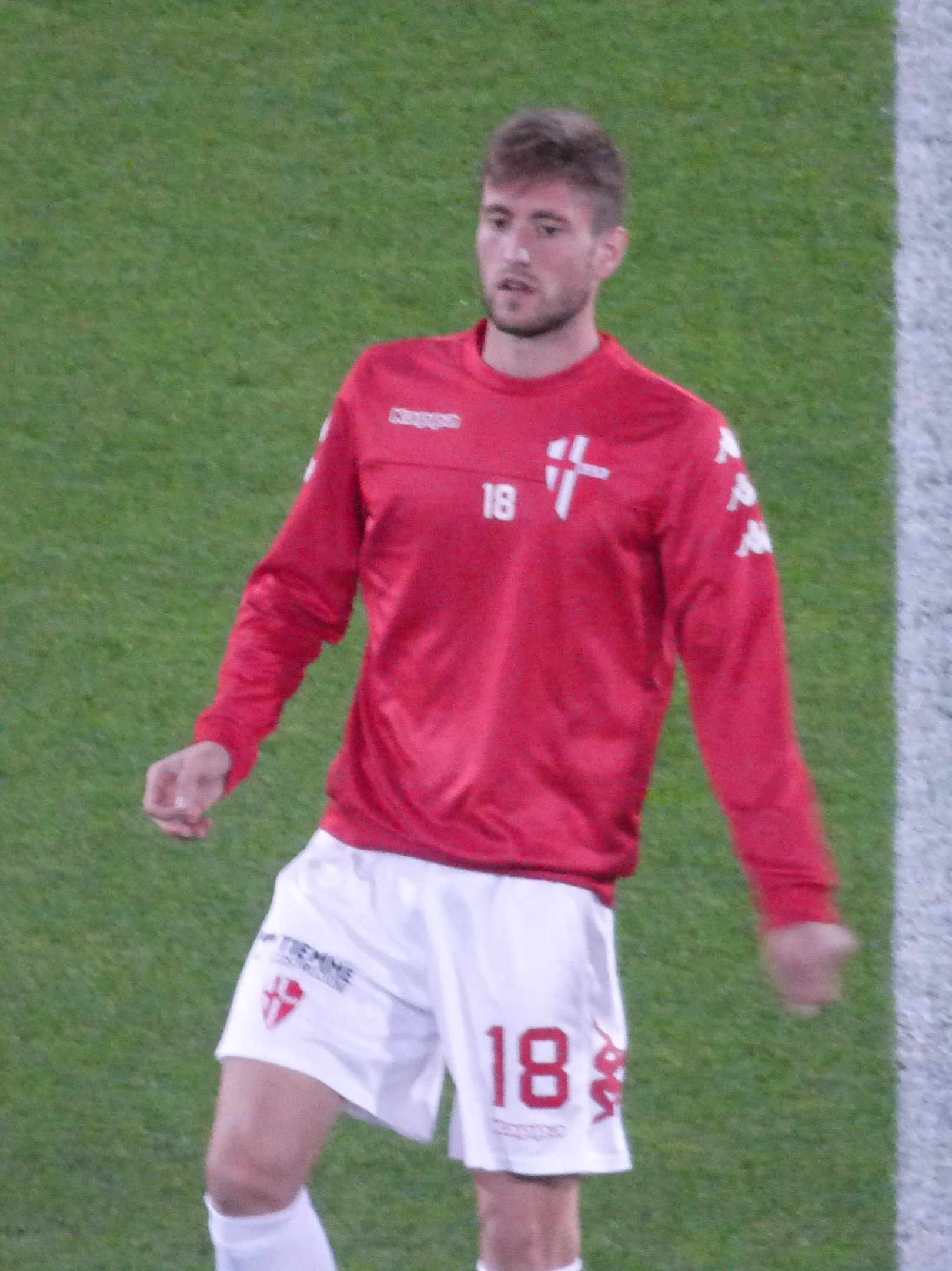 Ein Fußballspieler in rotem T-Shirt und weißen Shorts steht auf einem Feld mit ausgestreckten Armen und schaut nach vorne.