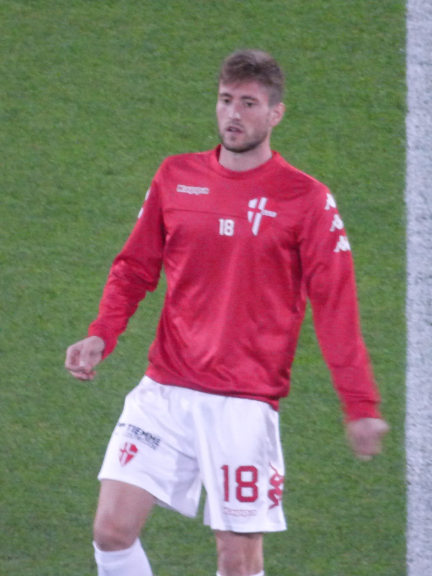 Ein Fußballspieler in rotem T-Shirt und weißen Shorts steht auf einem Feld mit ausgestreckten Armen und schaut nach vorne.