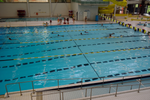 Großes Hallenbad mit schwimmenden Menschen, umgeben von Geländern und einem gefliesten Boden, Glasfenster an der Rückwand und Gegenstände auf der rechten Seite.