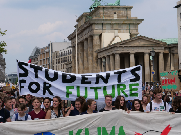 Eine Gruppe von Studenten marschiert in Berlin, die eine bunt bemalte Fahne mit der Aufschrift "Students for Future" schwenken, mit Gebäuden, Bäumen und Himmel im Hintergrund.