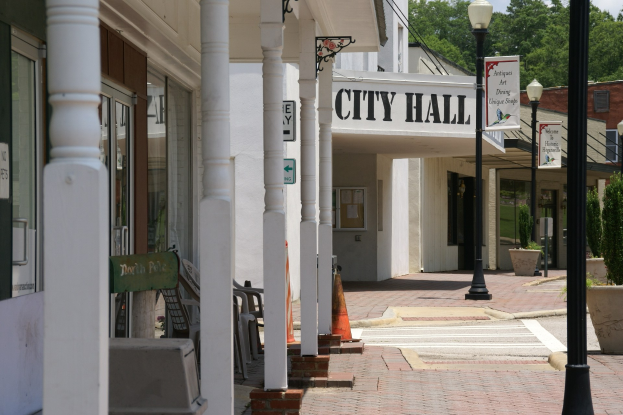 Rathaus mit umliegenden Gebäuden, Straßeninfrastruktur, Kraftfahrzeugen, Absperrgittern, Stühlen, Topfpflanzen, Bäumen und einem bewölkten Himmel.