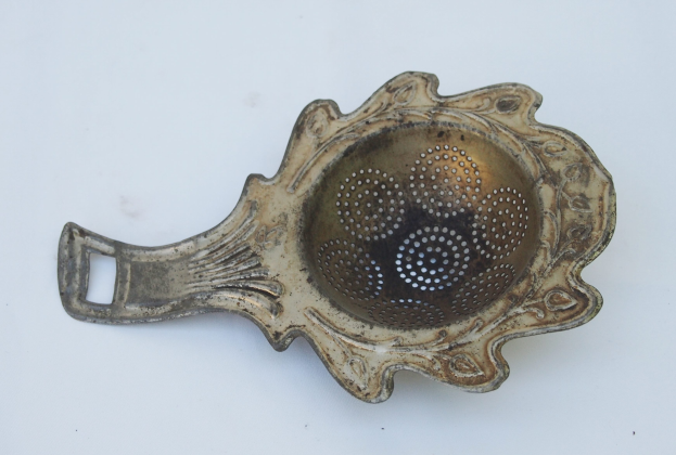 Ein Metallsieber mit einem Griff auf einer Seite und einem Ausguss auf der anderen, das eine gebogene Form mit einem zentralen Loch und einer kleinen oberen Öffnung hat, steht auf einer weißen Oberfläche.