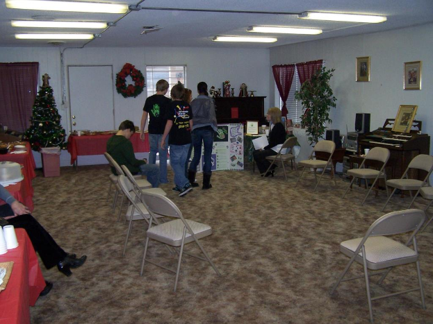 Ein Raum mit mehreren Stühlen, Tischen und Menschen, einige stehen, einige sitzen. Auf den Tischen stehen Becher und Gefäße. Im Hintergrund ist eine Wand mit einem Strauß, Vorhang, Tür, Fotorahmen und Fenstern zu sehen. Ein Weihnachtsbaum und ein Piano sind zu erkennen, mit Röhrenleuchten an der Decke.