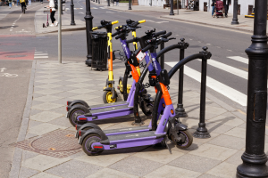 Eine Reihe von Elektro-Scootern, die auf der Straße geparkt sind, mit Menschen, die auf dem Gehweg laufen und Fahrräder fahren, einem Müllcontainer im Vordergrund und Gebäuden, Bäumen und Pflanzen im Hintergrund.