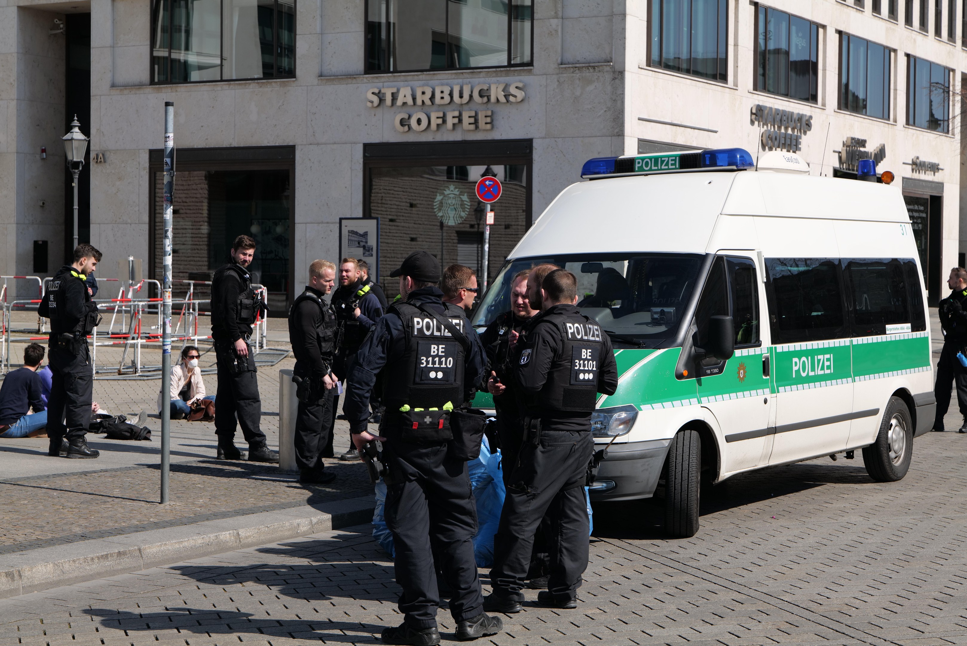 Eine Gruppe von Polizisten steht vor einem Starbucks-Café, rechts davon parkt ein Van und links stehen und sitzen ein paar Menschen; im Hintergrund ist ein Gebäude mit Fenstern, ein Schild, ein Laternenmast und ein Zaun zu sehen.