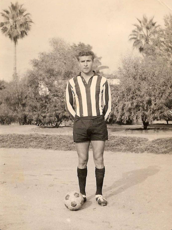 Ein Schwarz-Weiß-Foto eines alten Mannes, der einen Fußball hält und vor Bäumen und Himmel steht.