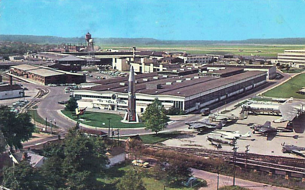 Altes Schwarz-Weiß-Foto eines Flughafens mit geparkten Flugzeugen, Gebäuden, Bäumen, Straßen, Fahrzeugen, Strommasten, Gras, Bahnschienen, Türmen, Hügeln und einem klaren blauen Himmel mit Text unten.