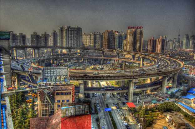 Stadtpanorama mit einer zentralen Ringstraße mit Fahrzeugen, einer unteren Straße mit Autos und Fahrrädern, zahlreichen Hochhäusern im Hintergrund, einem Himmel oben, Bäumen links unten und kleinen Häusern verteilt in der Stadt.