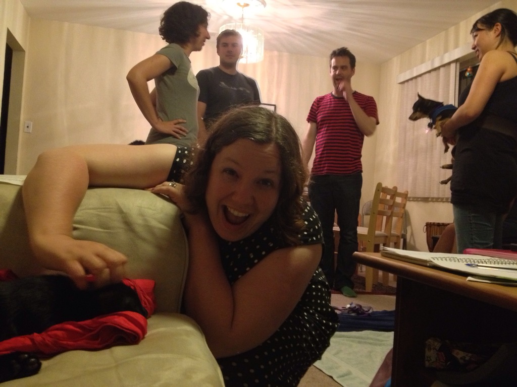 Eine Gruppe von fünf Menschen, drei Frauen und zwei Männer, in einem hell erleuchteten Raum mit einem Kronleuchter; eine Frau beugt sich lachend auf einem Sofa nach unten, eine andere hält einen Hund und trägt eine schwarze Bluse mit Jeanshosen, die dritte Frau hat kurze Haare und hellen Teint; die beiden Männer sprechen mit der Frau mit kurzen Haaren; es gibt zwei Stühle in der Ecke und einen Tisch auf der rechten Seite des Raums.