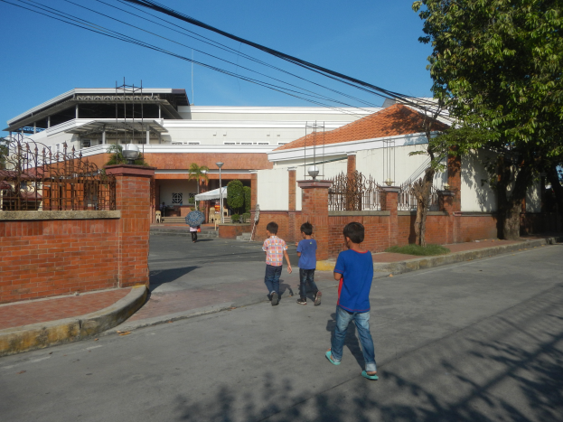 Eine Gruppe von Kindern geht auf einer Straße vor einem Gebäude mit Fenstern, einem Metallzaun, einem Tor, einigen Pflanzen, Gras, einem Straßenschild, einigen Bäumen, Drähten und einem bewölkten Himmel.