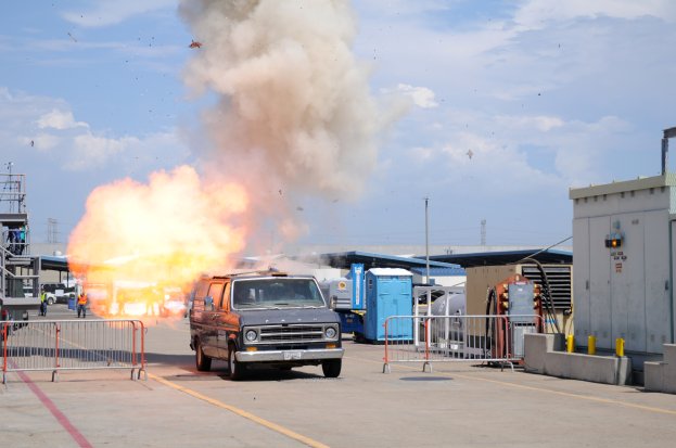 Ein Lieferwagen fährt eine Straße entlang mit einer großen Explosion daraus, umgeben von Absperrungen und anderen Fahrzeugen, mit einem Gebäude auf der rechten Seite und Schuppen, Pfählen und einem bewölkten Himmel im Hintergrund.