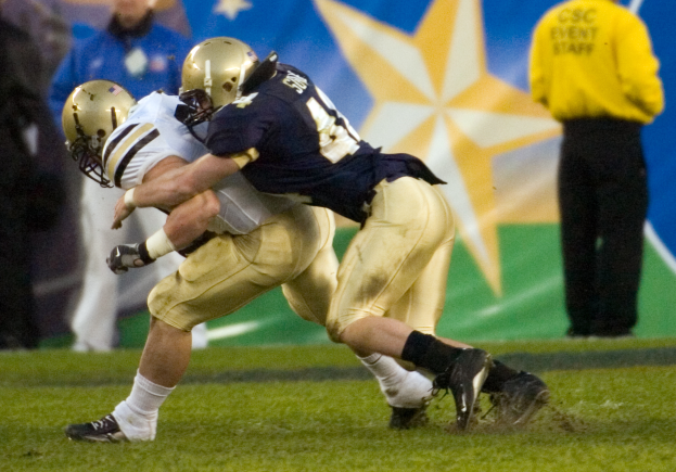 Ein Fußballer in Helm und Sportkleidung wird von einem anderen Spieler auf einem Rasenplatz getackelt, im Hintergrund sind einige Menschen und eine Fahne zu sehen.