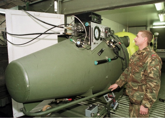 Ein Mann in militärischer Uniform steht neben einer großen grünen Bombe in einem unordentlichen Raum mit Maschinen, Kabeln und beleuchteten Wänden, mit Text unten.