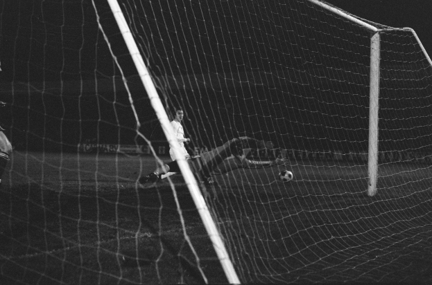 Ein Fußballer in einem Schwarz-Weiß-Foto hält den Ball, mit einem Ball in der Luft und einem Netz im Hintergrund.