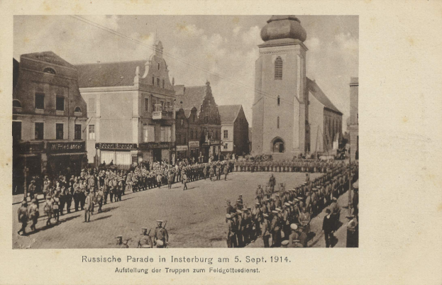 Ein Schwarz-Weiß-Foto eines Umzugs in Insterburg am 5. September 1914 mit vielen Menschen, Gebäuden und bewölktem Himmel sowie einem Text am unteren Rand.