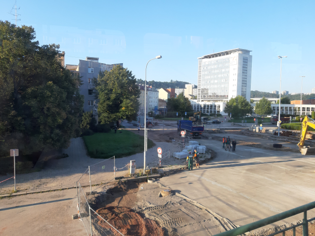 Eine Baustelle mit einem großen Loch, umgeben von einem Metallzaun, mit einer Gruppe von Menschen, Fahrzeugen, Pfählen, Schildern, einem Zaun, Gras, Pflanzen, Bäumen, Straßenlaternen, einem Turm, Gebäuden mit Fenstern und einem bewölkten Himmel.