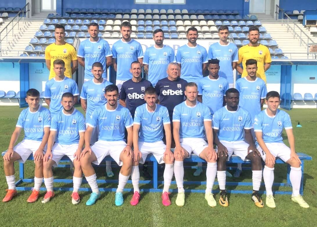 Gruppe von Männern, einige sitzen auf Bänken und einige stehen auf einem Fußballfeld, mit Stühlen, Treppen, Geländern, Wänden und Fenstern im Hintergrund.