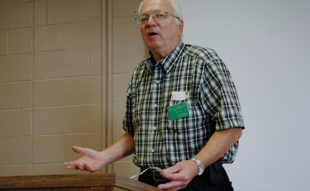 Eine Person steht an einem Podium, hält ein Papier und hat eine Wand und eine Tafel im Hintergrund.