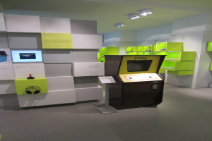 Ein hell erleuchteter Raum voller verschiedener Videospiele, mit einem zentralen Arcade-Automaten, einem Display mit Text auf der linken Seite und Regalen mit Gegenständen auf der rechten Seite.