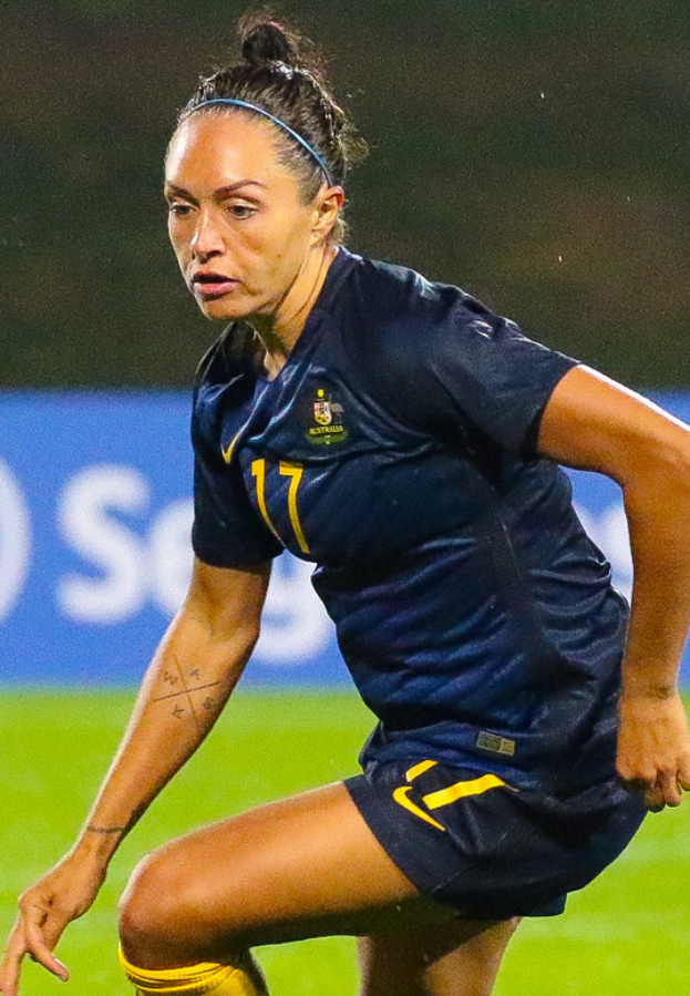 Eine Fußballspielerin in Aktion auf dem Feld, die ein blaues T-Shirt und gelbe Shorts trägt, mit einem unscharfen Hintergrund, der ein blaues Schild mit Text zeigt.