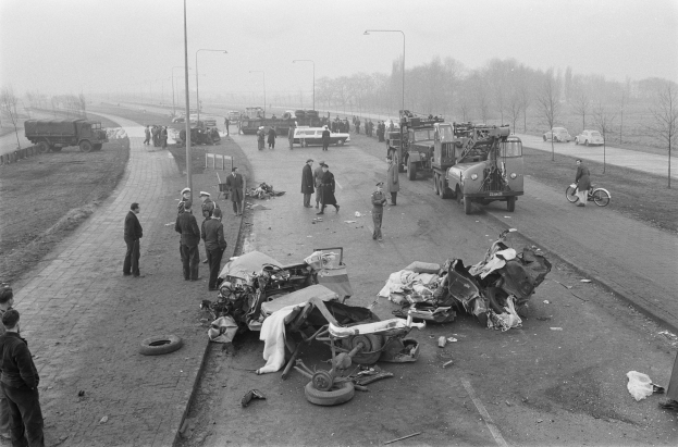 Schwarz-weißes Bild einer Autounfall-Szene am Straßenrand mit mehreren Fahrzeugen, darunter ein Landfahrzeug, und einer Gruppe von Menschen drumherum, mit Laternenpfählen, Bäumen und Himmel im Hintergrund.