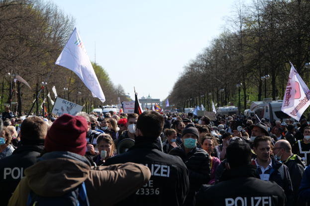 Eine große Gruppe von Menschen steht vor einer Menge von Polizeibeamten, einige tragen Mönen und Masken, während einer Demonstration in Berlin, Deutschland, mit Schildern, Fahnen, Laternenmästen, Bäumen, Fahrzeugen, einem Gebäude und dem Himmel im Hintergrund.