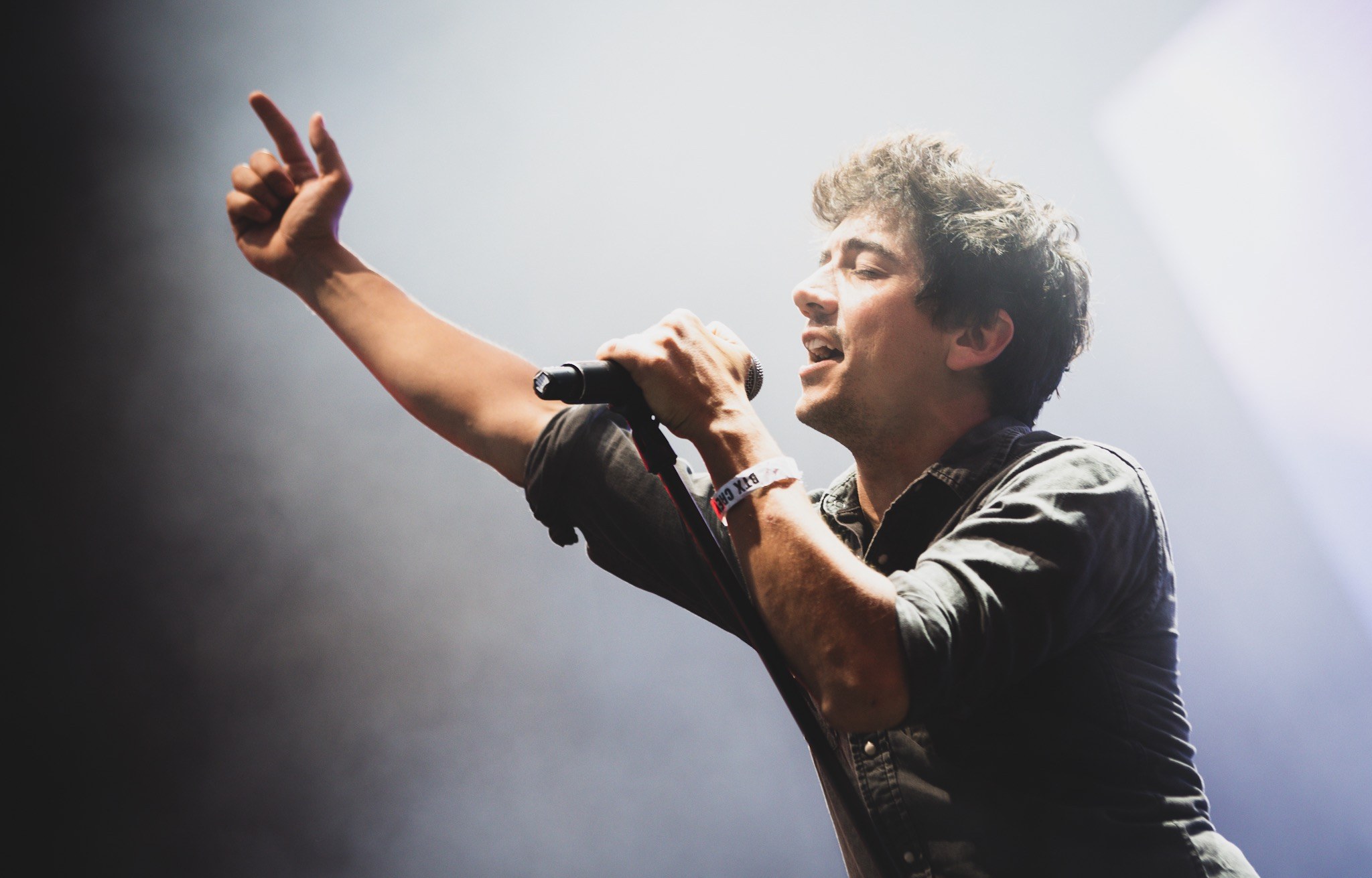 Ein Mann in schwarzem Hemd und Jeans singt leidenschaftlich in ein Mikrofon auf einer hell erleuchteten Bühne, seine Augen geschlossen und das Haar stylisch durcheinander, während das Publikum im Hintergrund applaudiert.