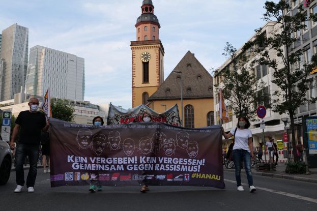 Eine Gruppe von Menschen mit Masken, die eine Plakette tragen und auf der Straße gehen, mit einem geparkten Auto auf der linken Seite, Gebäude und Bäume im Hintergrund und einem klaren blauen Himmel.