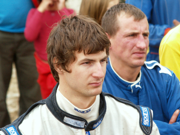 Eine Gruppe von Menschen auf dem Boden, mit einem Mann in einem Rennanzug neben einem anderen Mann sitzend, die Arme verschränkt und einen entschlossenen Gesichtsausdruck.