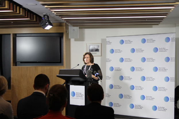 Eine Frau steht auf einer B├╝hne und spricht in ein Mikrofon, mit einem Publikum vor ihr, einem Banner an der Wand hinter ihr, einem Fernseher an der Holzwand und Lichtern an der Decke.