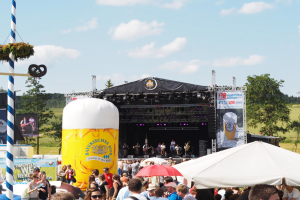 Große Menschenmenge beim Oktoberfest in München, Deutschland, vor einer Bühne mit Musikern, viele tragen Mützen und Brillen, einige halten Schirme, mit einem dekorierten Pfahl und einem Zelt im Vordergrund, Bäume und bewölkter Himmel im Hintergrund.