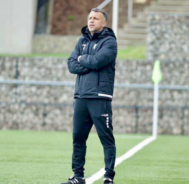 Ein Mann in Schwarz steht auf einem Fussballfeld mit überkreuzten Armen, scheinbar ein Trainer während eines Trainings.