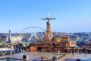 Ein belebter Stadtplatz mit einem zentralen Riesenrad, umgeben von Gebäuden, Bäumen und Menschen unter einem klaren blauen Himmel, mit Marktzelten und Geländern, mit einem Wasserzeichen, das darauf hinweist, dass es einer der besten Weihnachtsmärkte in Europa ist.