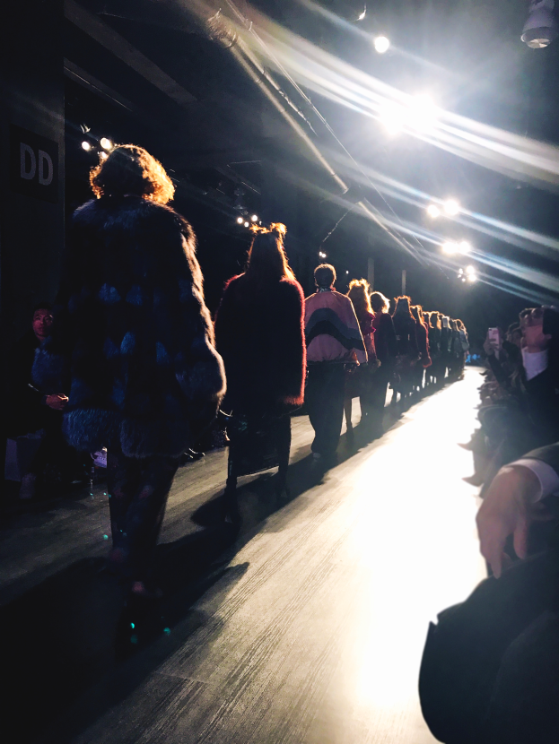 Eine Gruppe von Menschen läuft über den Laufsteg einer Modenschau, mit Zuschauern auf beiden Seiten.