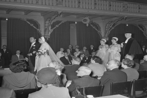 Ein Schwarz-Weiß-Foto einer Hochzeitszeremonie in einem großen Raum mit Sitzenden und Stehenden, einige halten Blumensträuße, und Tische mit Tassen und anderen Gegenständen, mit Vorhängen und Deckenleuchten im Hintergrund.