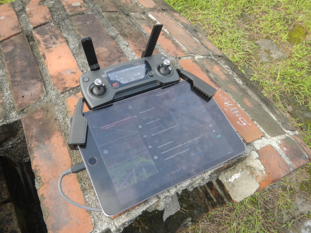 Ein Tablet-Computer auf einer Ziegelwand, verbunden durch ein Kabel mit einem DJI Mavic 2 Enterprise Fly More Combo, mit grünem Gras im Hintergrund.