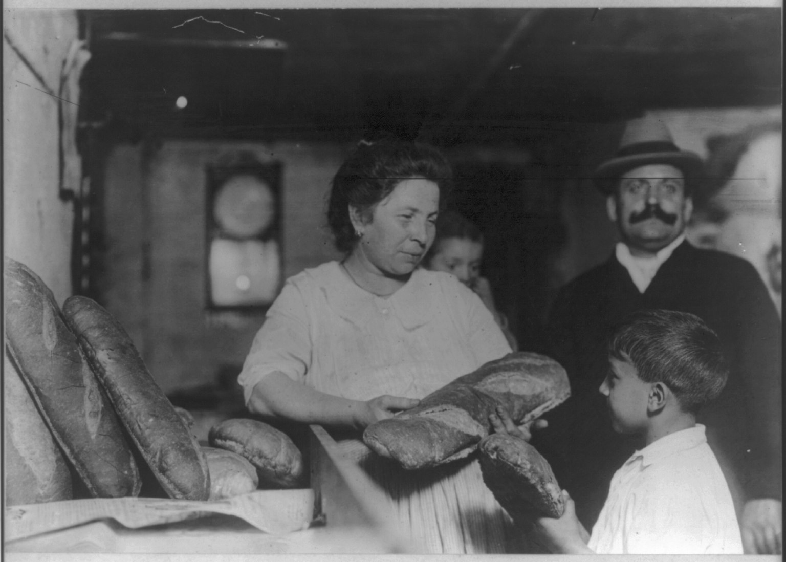 Altes Schwarz-Weiß-Foto einer Frau mit einem Brotlaib in der Hand und zwei Kindern neben ihr in einer Bäckerei, die andere Brote auf einem Tisch und ein gerahmtes Objekt an der Wand dahinter zeigt.