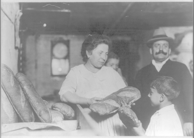 Altes Schwarz-Weiß-Foto einer Frau mit einem Brotlaib in der Hand und zwei Kindern neben ihr in einer Bäckerei, die andere Brote auf einem Tisch und ein gerahmtes Objekt an der Wand dahinter zeigt.
