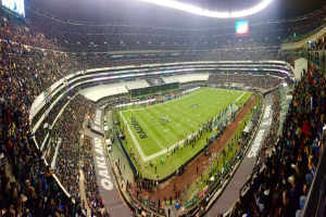 Ein großes Stadion mit Zuschauern, die ein Fußballspiel verfolgen, mit Spielern und Gegenständen auf dem Feld, beleuchtet von Deckenlampen, und mit Tafeln, Geländern und Bildschirmen.