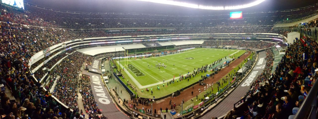 Ein großes Stadion mit Zuschauern, die ein Fußballspiel verfolgen, mit Spielern und Gegenständen auf dem Feld, beleuchtet von Deckenlampen, und mit Tafeln, Geländern und Bildschirmen.