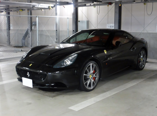 Schwarzer Ferrari California in einer Parkgarage geparkt, mit einer Wand im Hintergrund, Maschendraht auf der linken Seite und Deckenbeleuchtung.