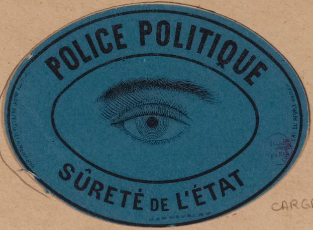 Ein brauner Karton mit einem blauen Aufkleber, auf dem "Polizei Politique Surete de L'Etat" steht und ein Auge in der Mitte.