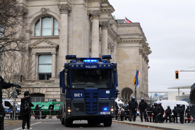 Gruppe von Polizisten vor einem großen Gebäude mit Fahrzeugen auf der Straße, eine Person mit einer Kamera auf der linken Seite, Bäume und Verkehrszeichen im Hintergrund und ein klarer blauer Himmel.