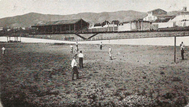 Ein Schwarz-Weiß-Foto einer Gruppe, die Cricket am Strand in den frühen 1900er Jahren spielt, mit Gebäuden, Bäumen, Hügeln und einer Brücke im Hintergrund und Text unten.